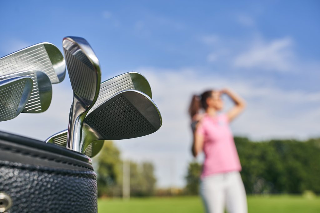 A close-up of the most forgiving golf irons nestled in a bag, with a woman in a pink shirt standing gracefully in the blurred background on a sunlit golf course.
